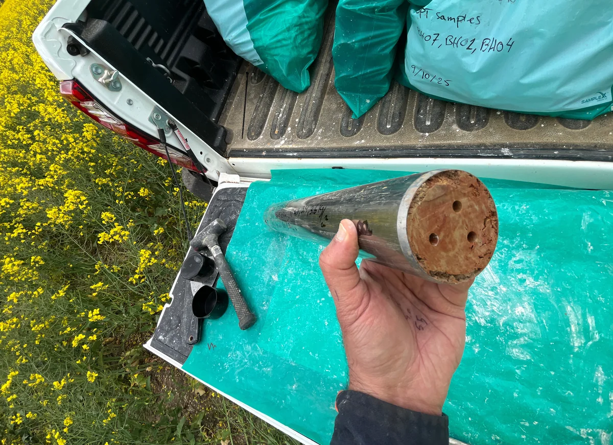 Recovered soil in a sampling tube held above a vehicle tailgate, with sample bags behind it at Joel Joel.